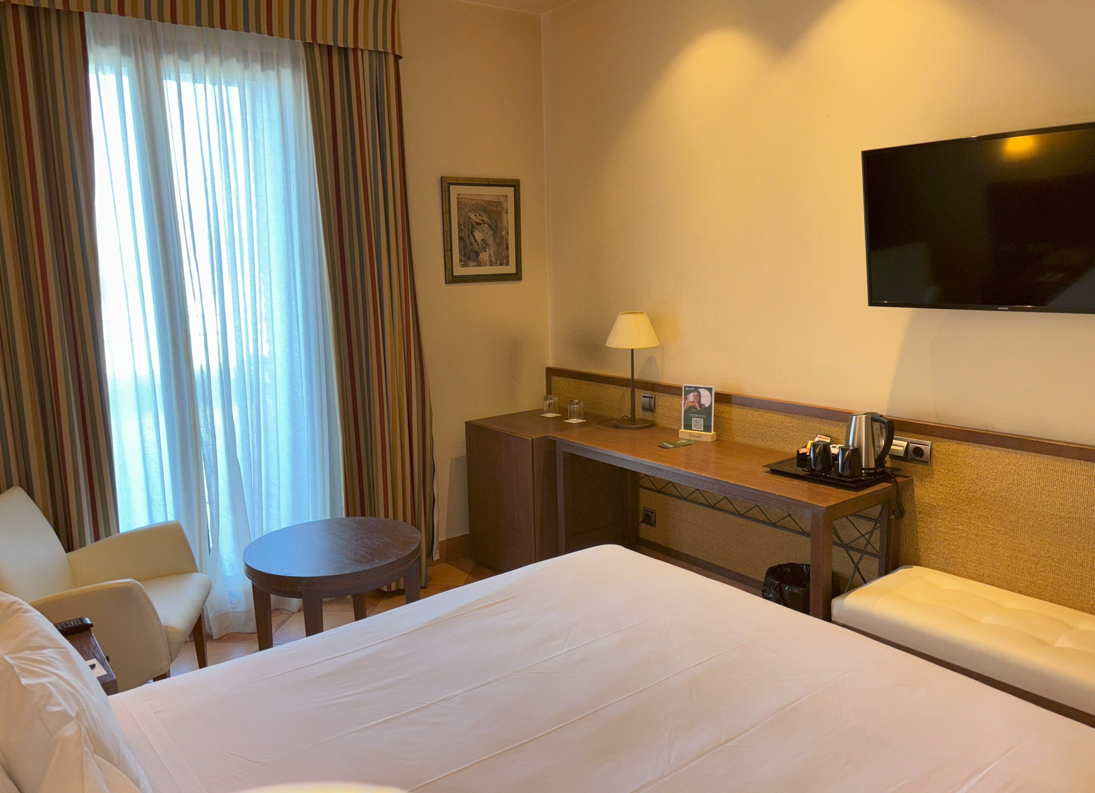 Habitación de hotel con una cama grande, un escritorio con lámpara, televisión y ventana con cortinas.