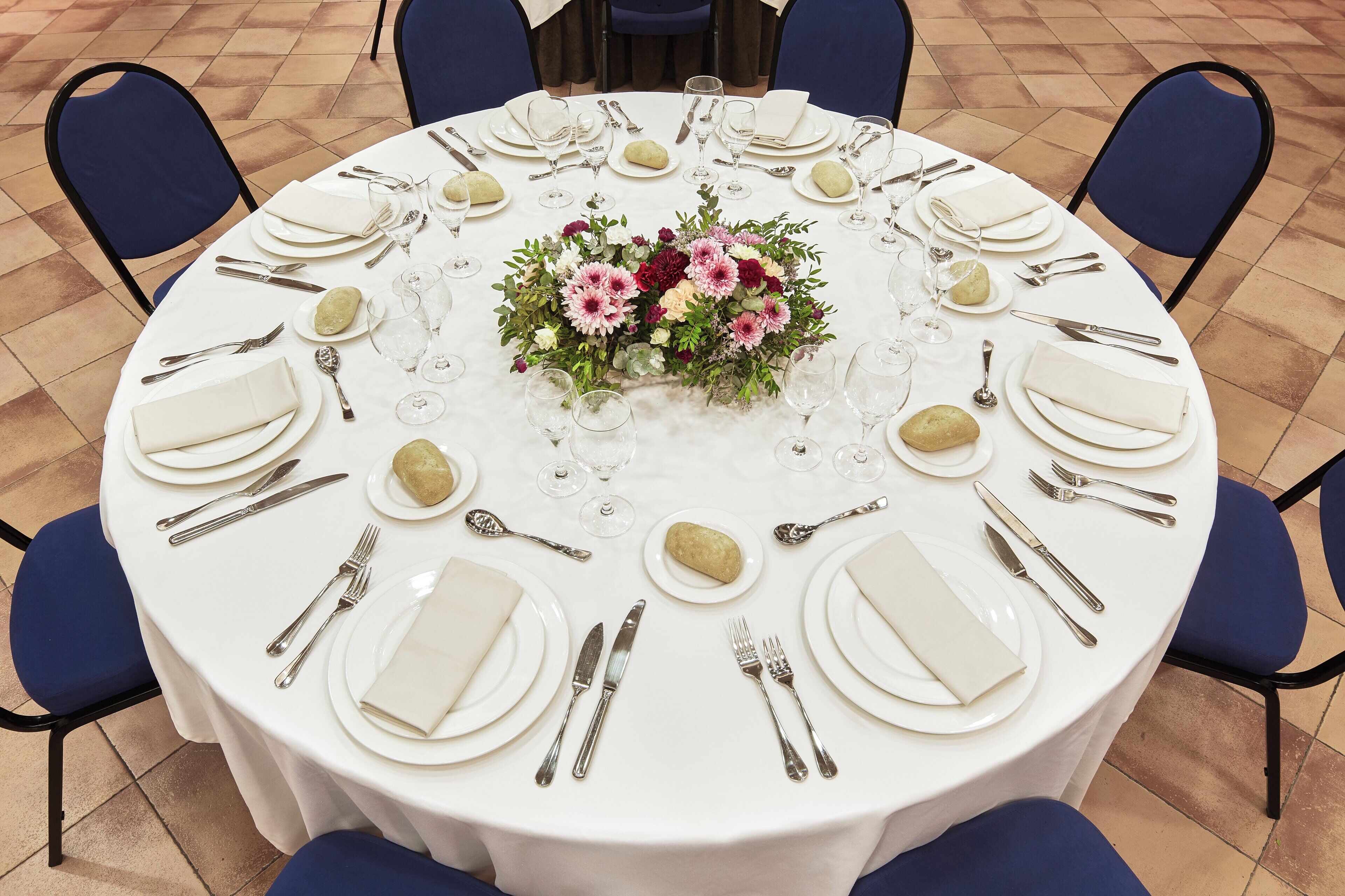Mesa redonda dispuesta para una cena formal, con mantel blanco, platos, cubiertos y un centro de mesa floral.