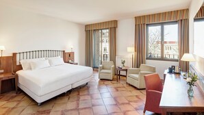 Habitación de hotel con una cama grande, dos sillones, una mesita y vistas a la ciudad a través de amplias ventanas.