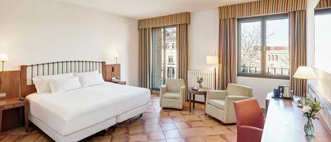 A hotel room with a large bed, two armchairs, a small table, and a view of the city through large windows.