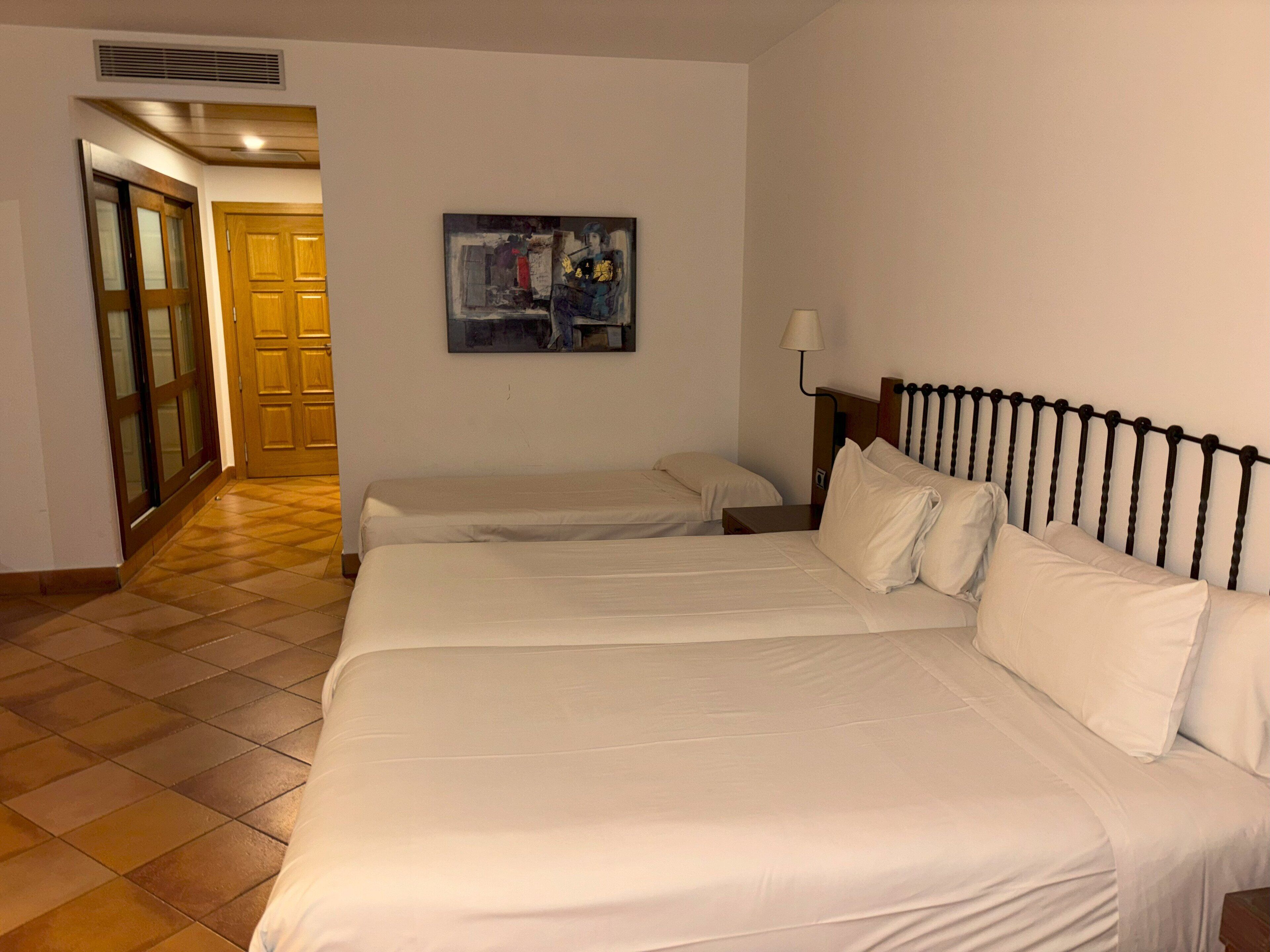 A hotel room with two beds, a painting on the wall, and a lamp.
