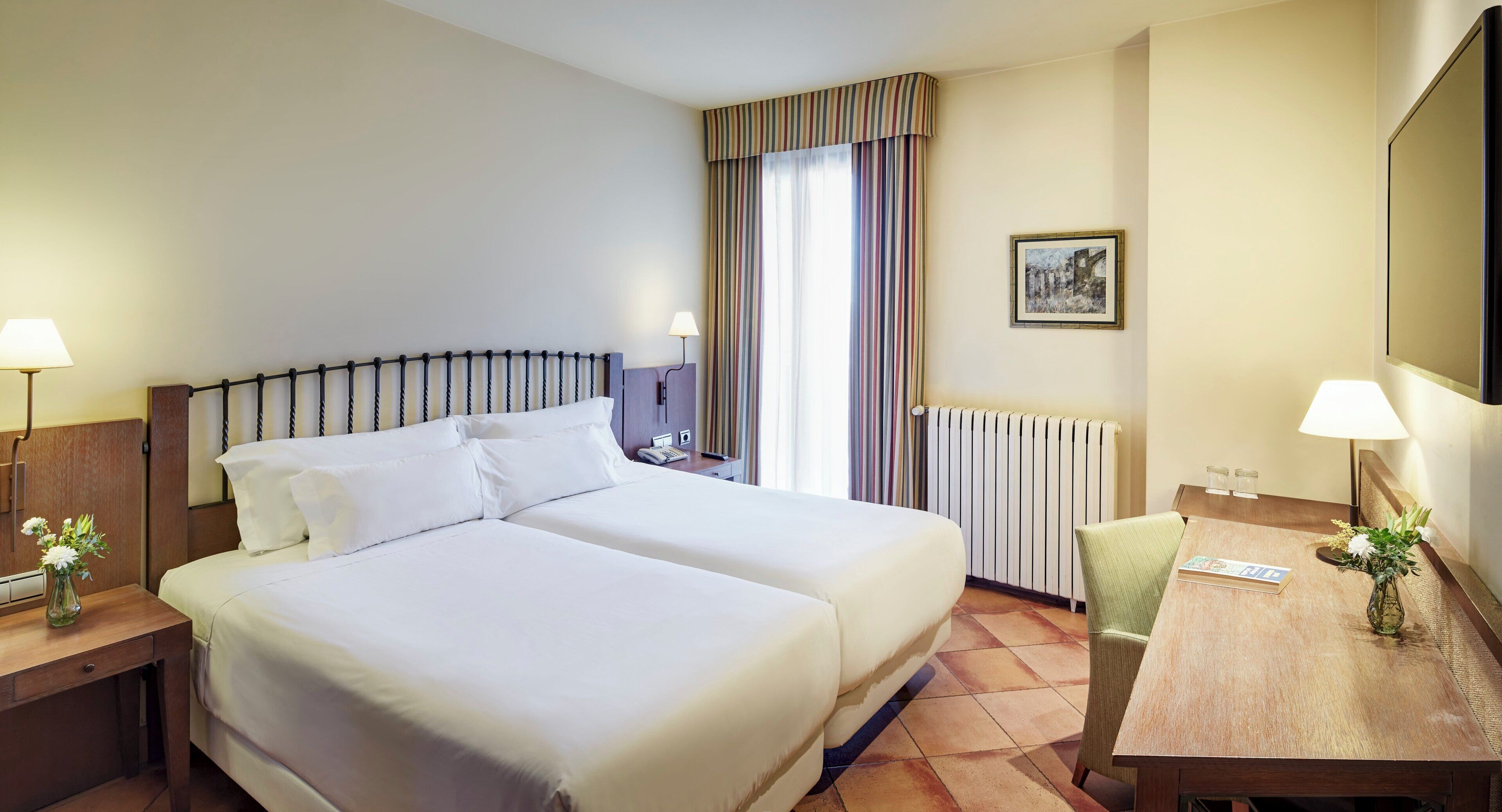Een hotelkamer met twee bedden, een bureau, een stoel, een radiator, een lamp, een foto aan de muur en een vaas met bloemen.