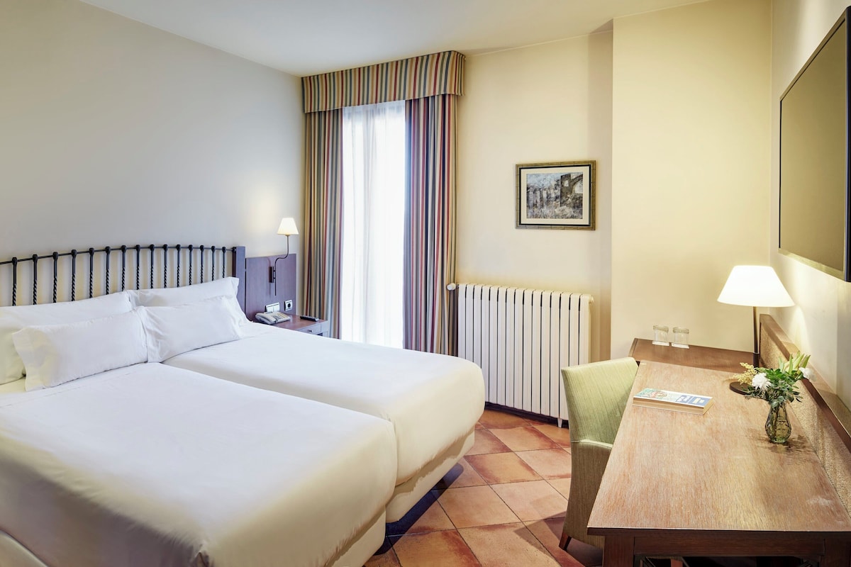 A hotel room with two beds, a desk, a chair, a radiator, a lamp, a picture on the wall, and a vase with flowers.