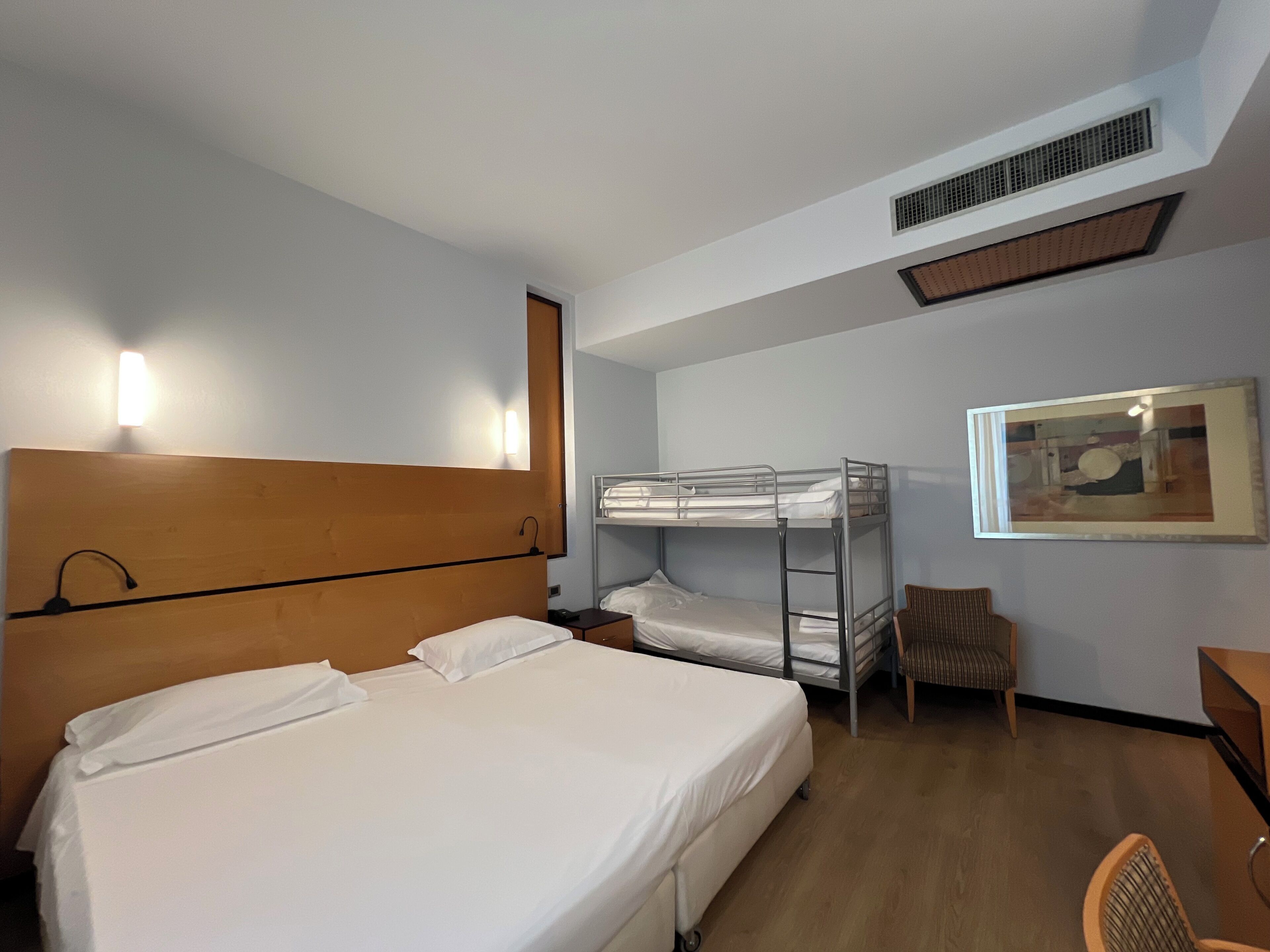 Una habitación de hotel con una cama grande, una cama más pequeña en la litera superior, una silla, un escritorio y un cuadro en la pared.