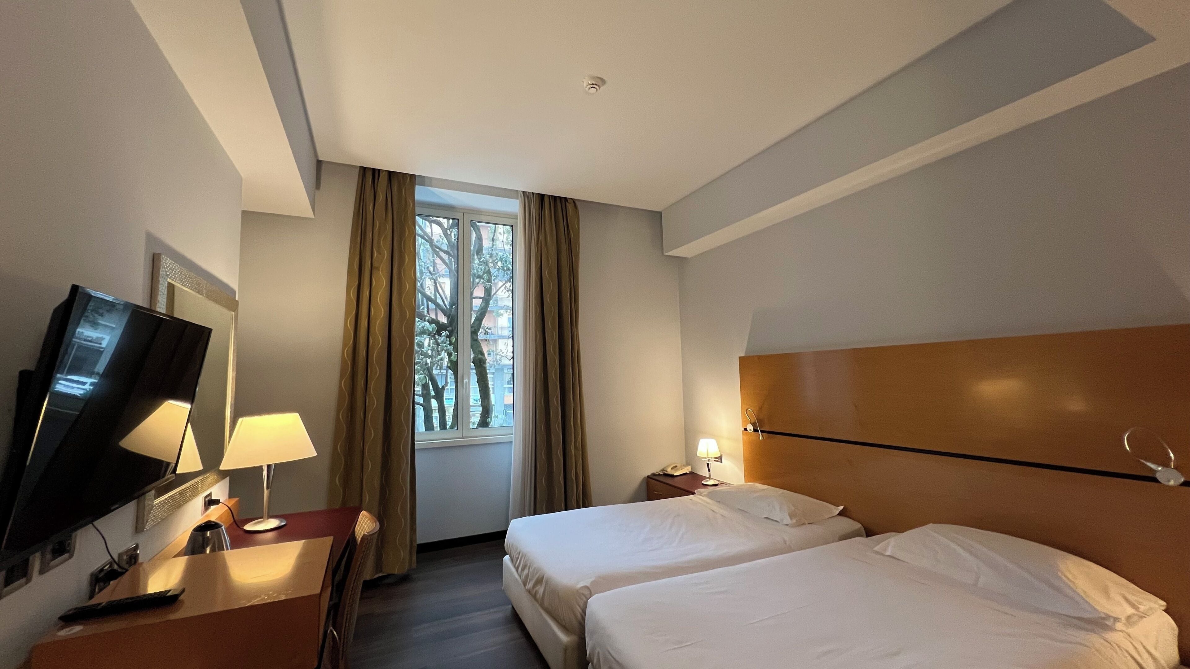 A hotel room with two beds, a TV, a desk with a lamp, a window with curtains, and a headboard.
