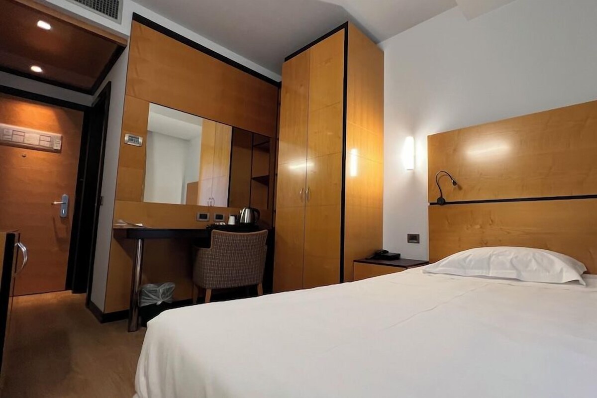 A hotel room with a bed, a desk, a chair, a wardrobe, a mirror, and a wall-mounted lamp.