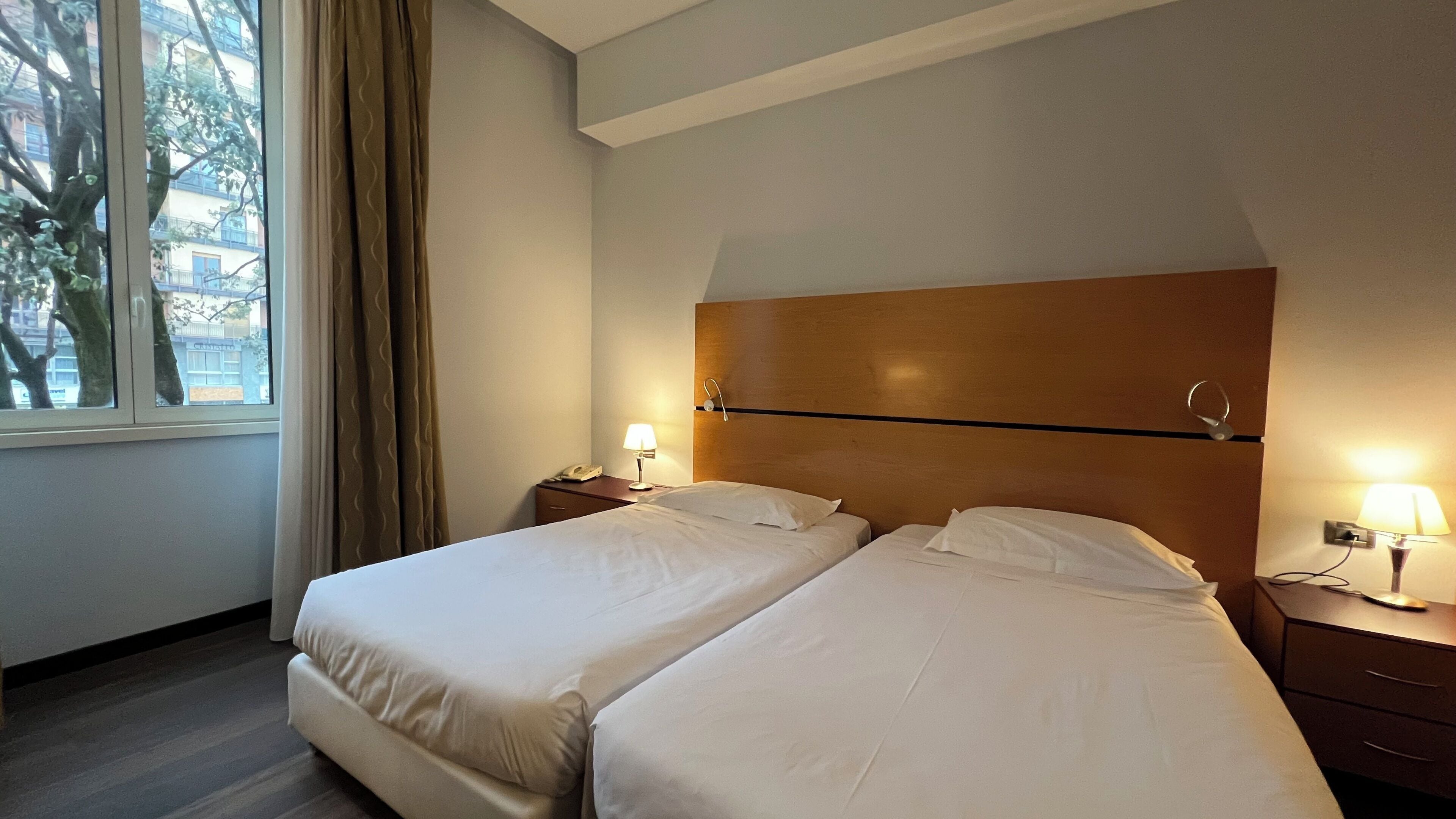Una habitación de hotel con dos camas, cabecero de madera, ventana con cortinas y lámparas de noche.