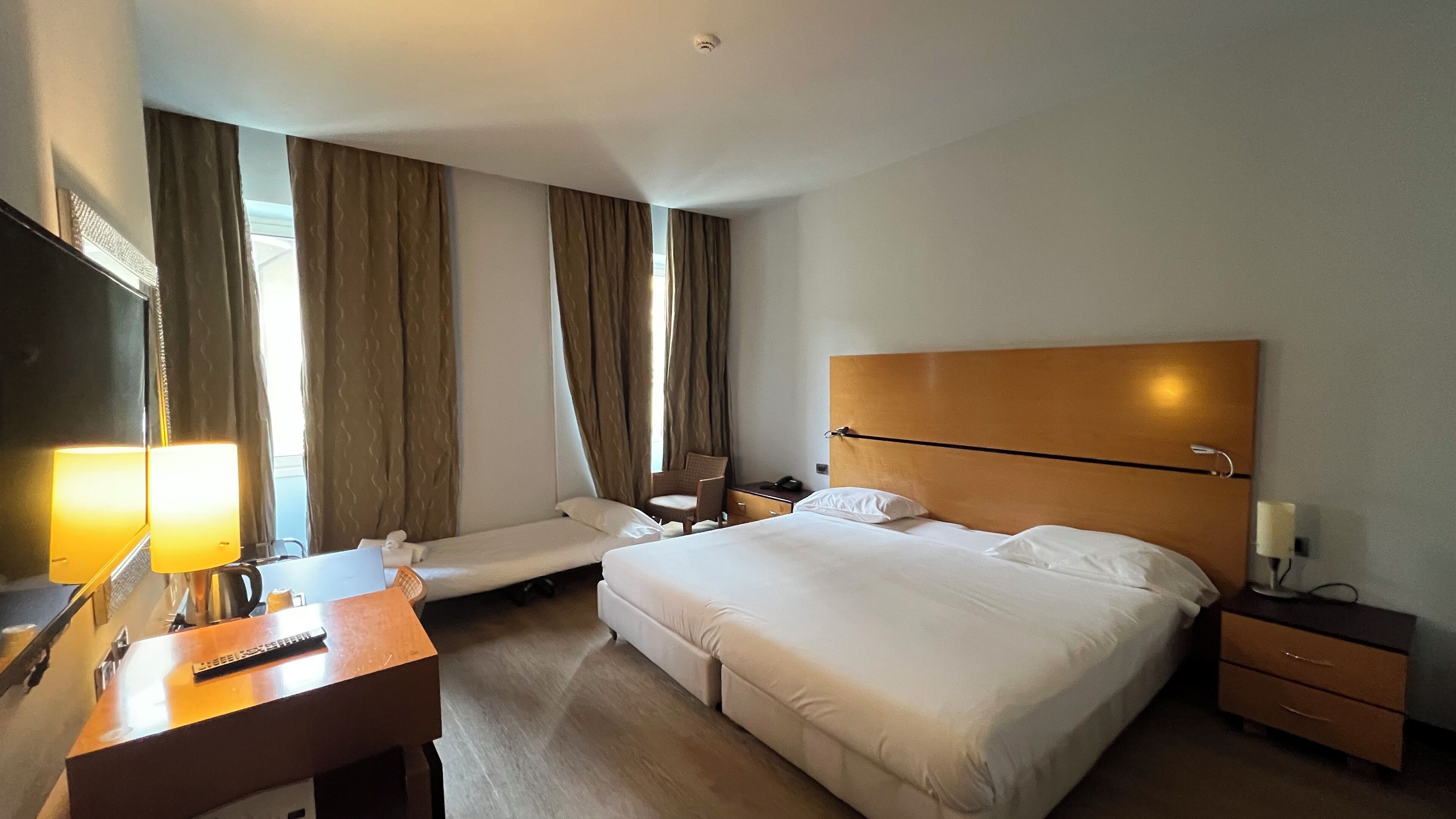 Ein Hotelzimmer mit Bett, Nachttischen, einem Schreibtisch mit Lampe, einem Sessel und einem Fernseher.