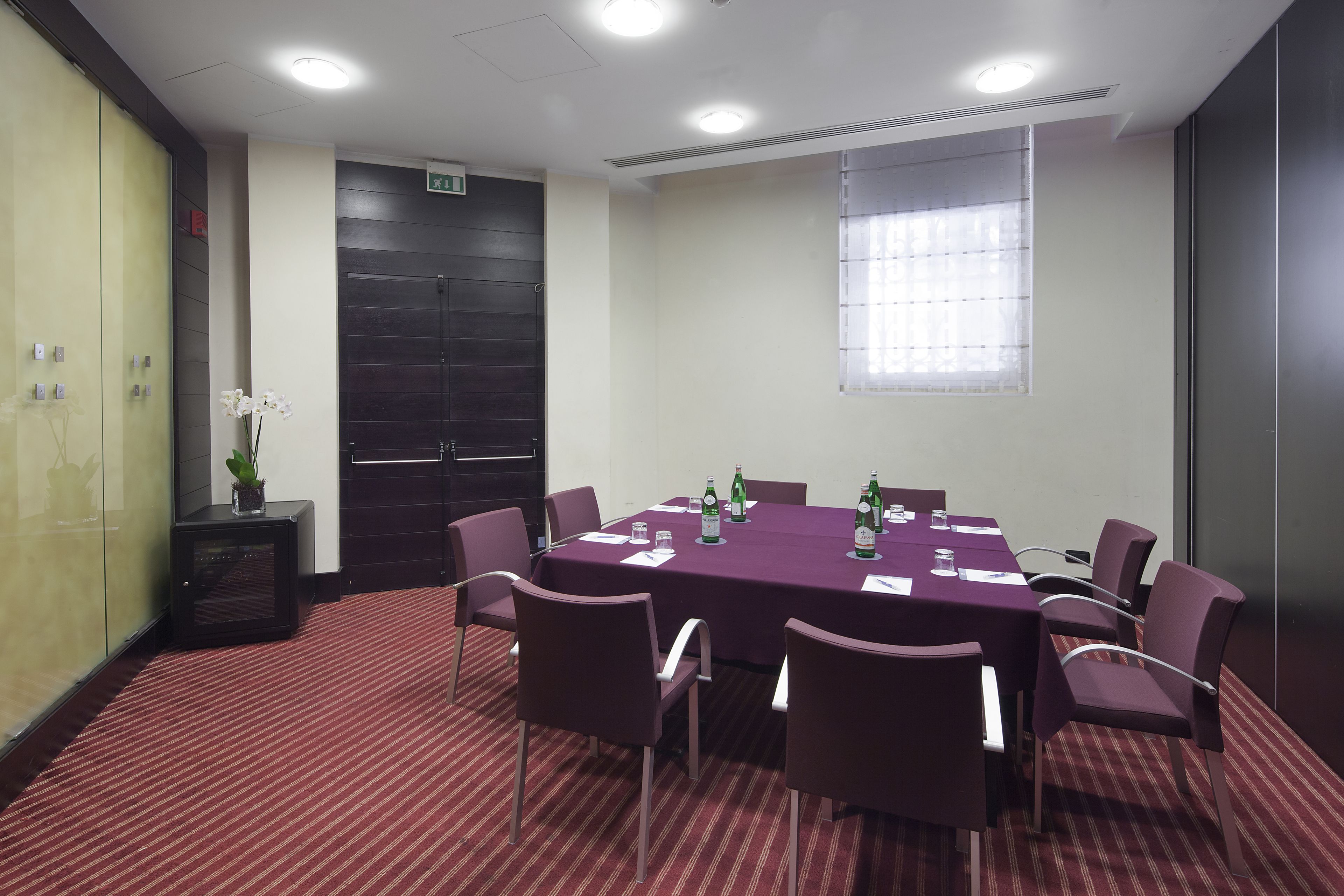 Un salón de conferencias con alfombra roja, sillas moradas y una mesa dispuesta con botellas y papeles.
