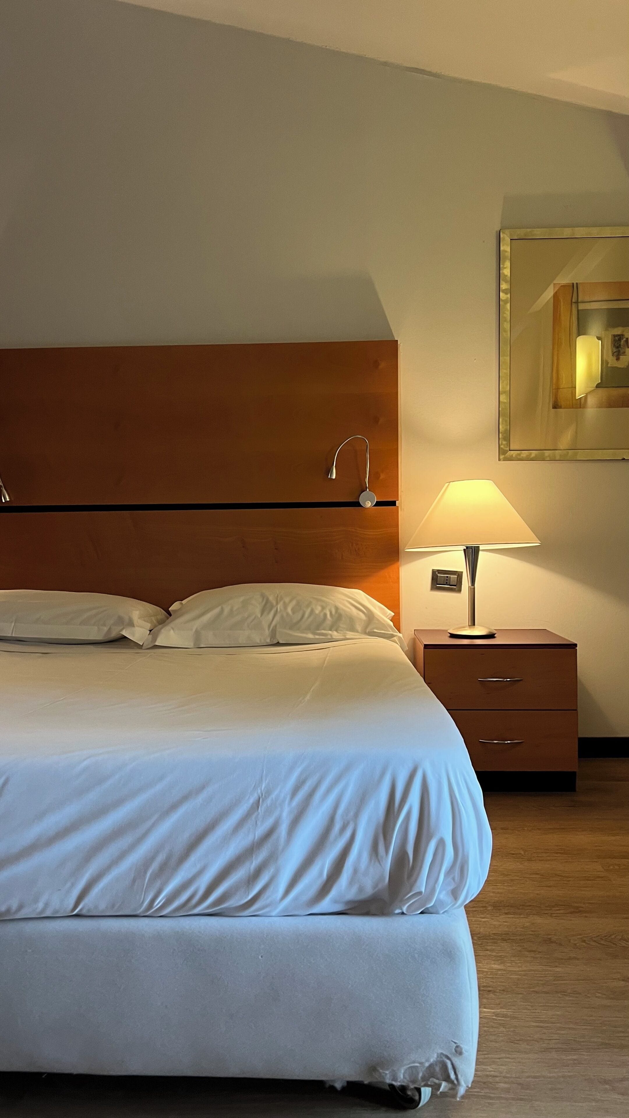 Una cama bien tendida con sábanas blancas, un cabecero de madera, una lámpara de noche, una mesita de noche con cajones y un espejo montado en la pared.