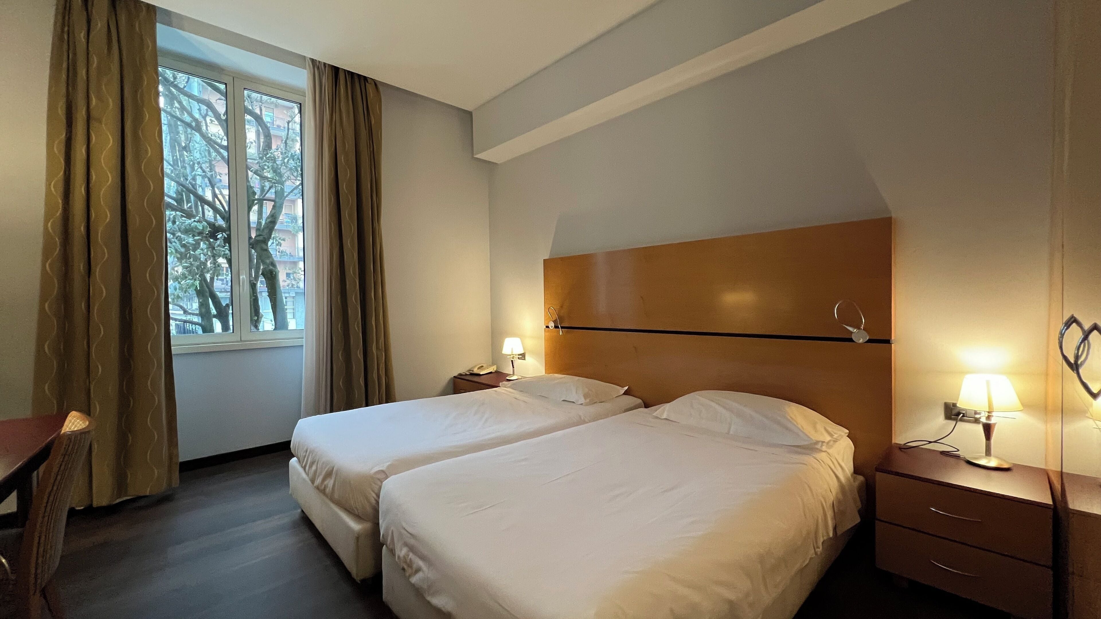 A hotel room with two beds, a desk, a chair, a nightstand with a lamp, and a window with curtains.