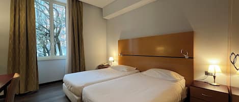 A hotel room with two beds, a desk, a chair, a nightstand with a lamp, and a window with curtains.