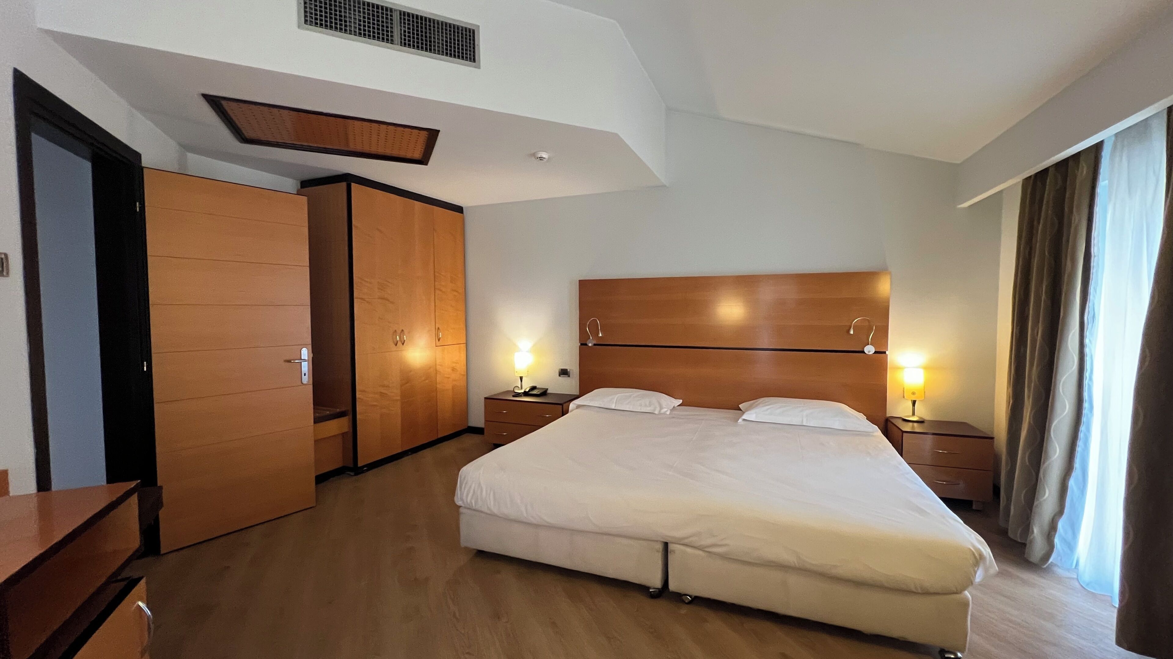 Habitación de hotel con una cama grande, armario de madera, mesitas de noche con lámparas y un respiradero en el techo.
