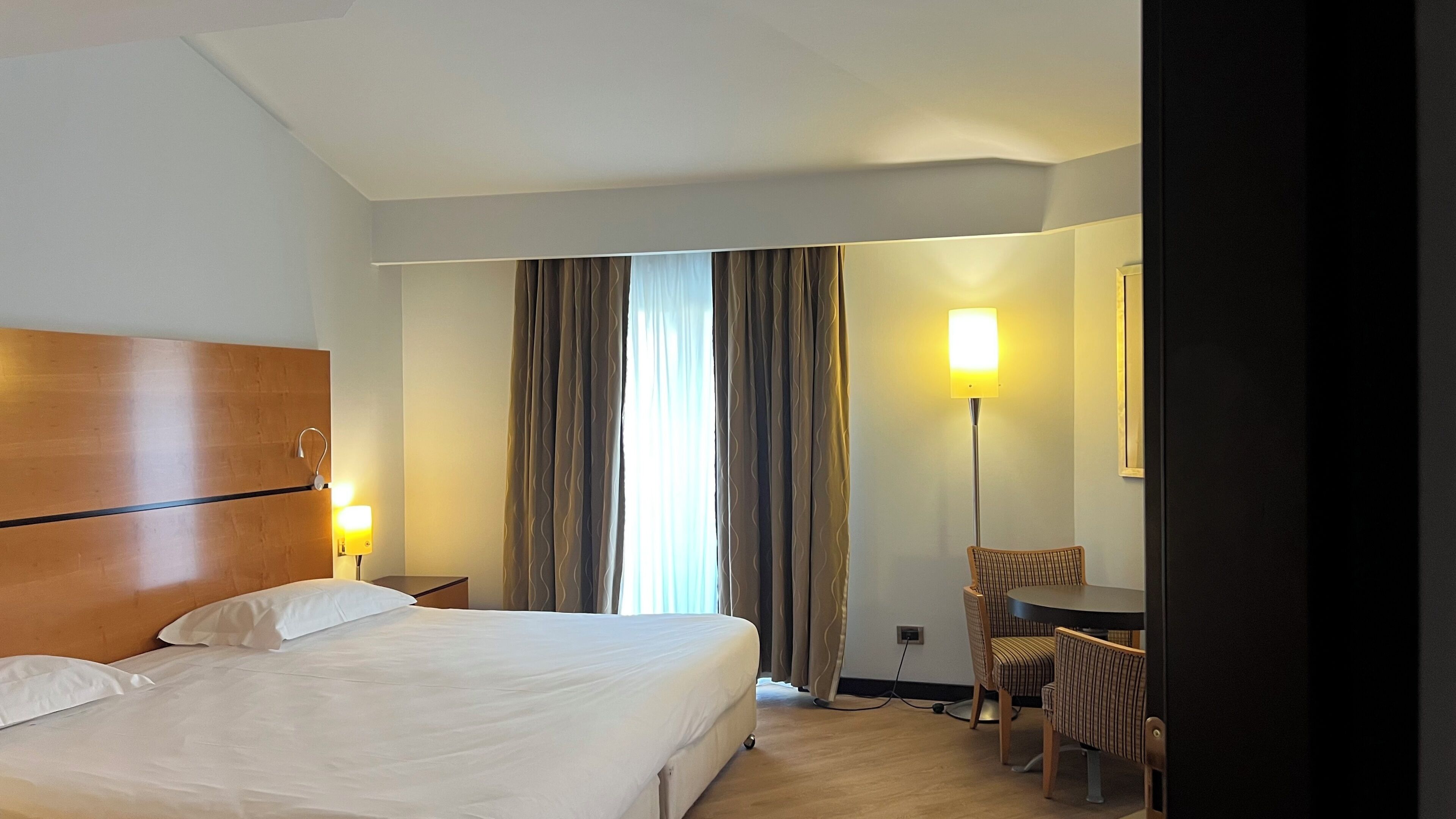 Una habitación de hotel con cama, escritorio, silla, lámpara y cortinas.