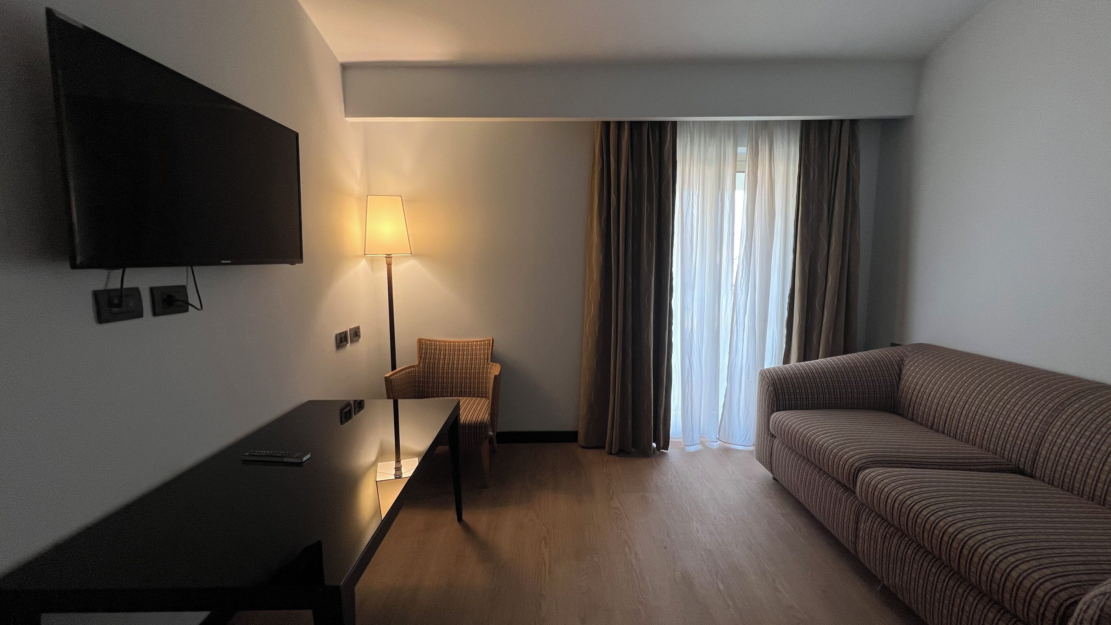 A hotel room with a TV mounted on the wall, a desk with a lamp, a striped sofa, and a chair.