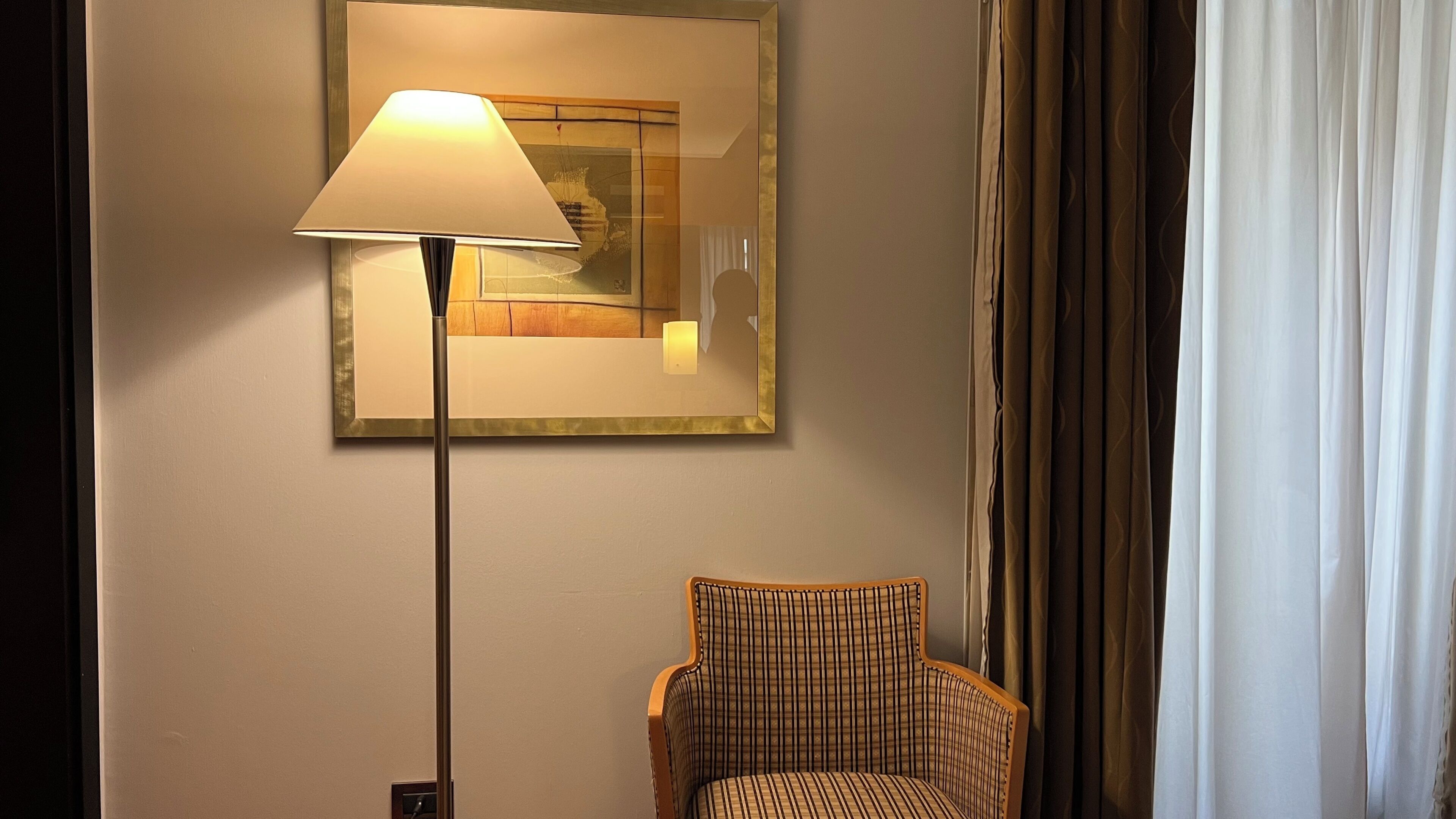 A room with a standing floor lamp, a framed picture on the wall, a plaid chair, and a curtain.
