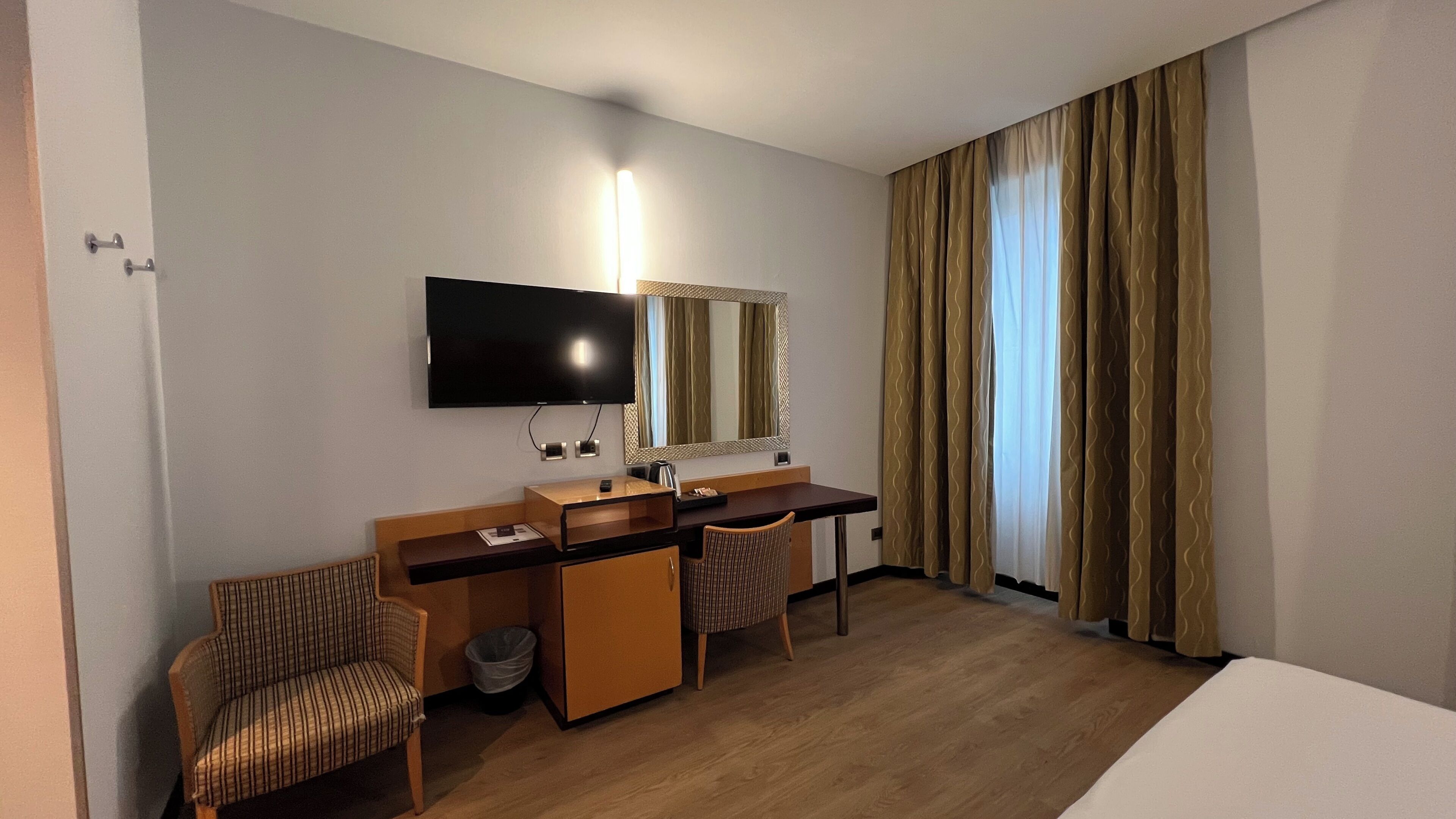 Une chambre d’hôtel comprenant un lit, un bureau avec une chaise, une télévision, un miroir et une fenêtre avec des rideaux.