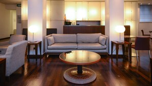 A modern living room with a round glass coffee table, a grey sofa, and two armchairs. There are two small side tables with lamps. The room has wooden flooring and a large window in the background. - Hotel Igea (Brescia)