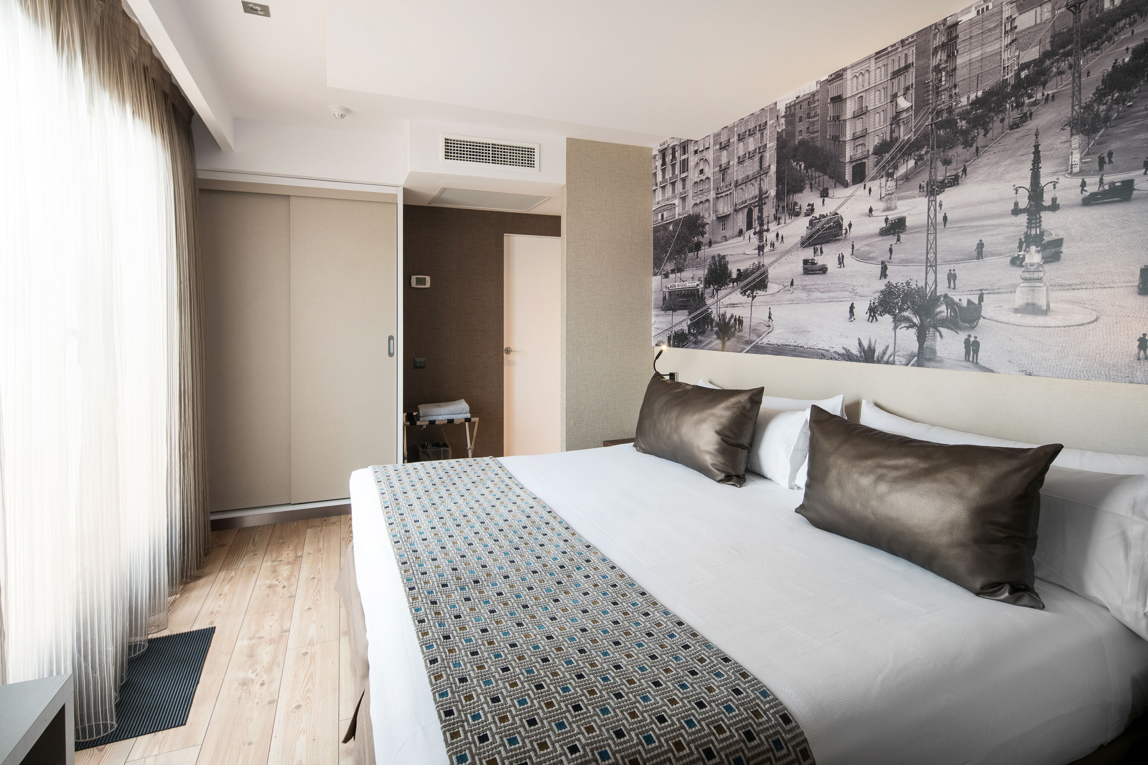 A hotel room with a large bed, a view of a cityscape through a window, and a door leading to a bathroom.