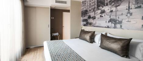 A hotel room with a large bed, a view of a cityscape through a window, and a door leading to a bathroom.