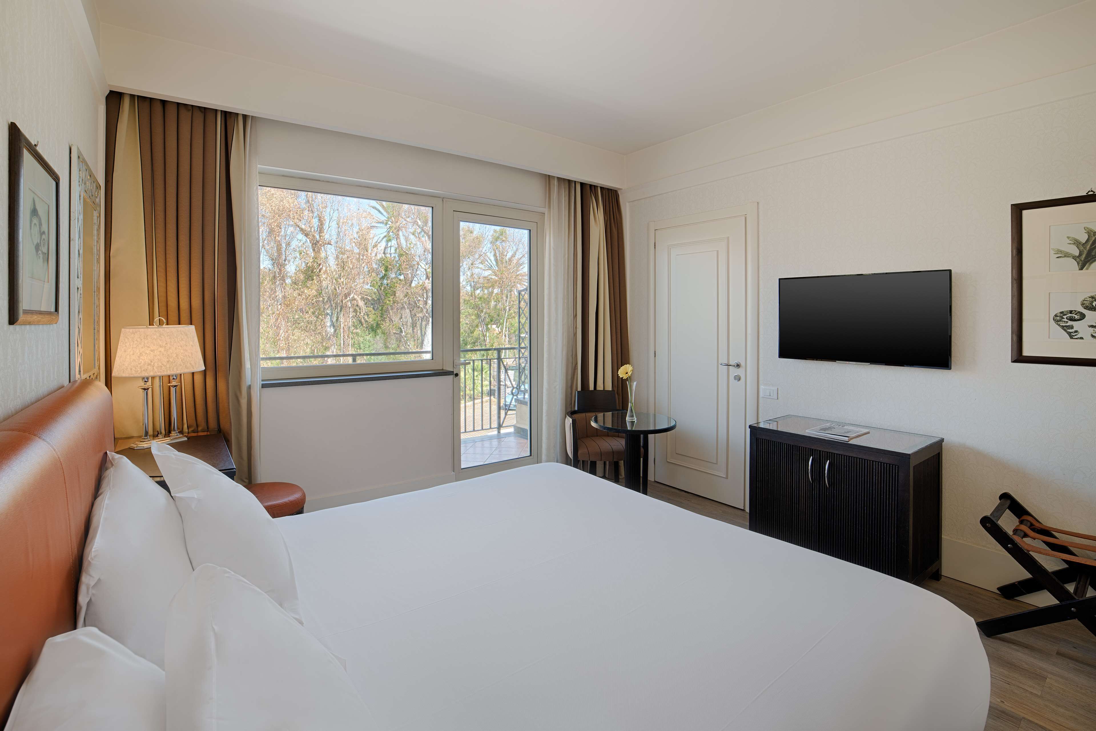 A hotel room with a large bed, a TV, a small table with a lamp, a window with curtains, and a balcony.