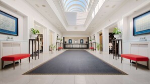 A spacious hotel lobby with a central reception desk, red benches, potted plants, and a skylight.