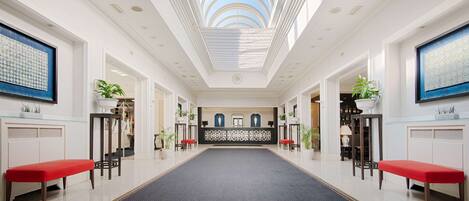 A spacious hotel lobby with a central reception desk, red benches, potted plants, and a skylight.