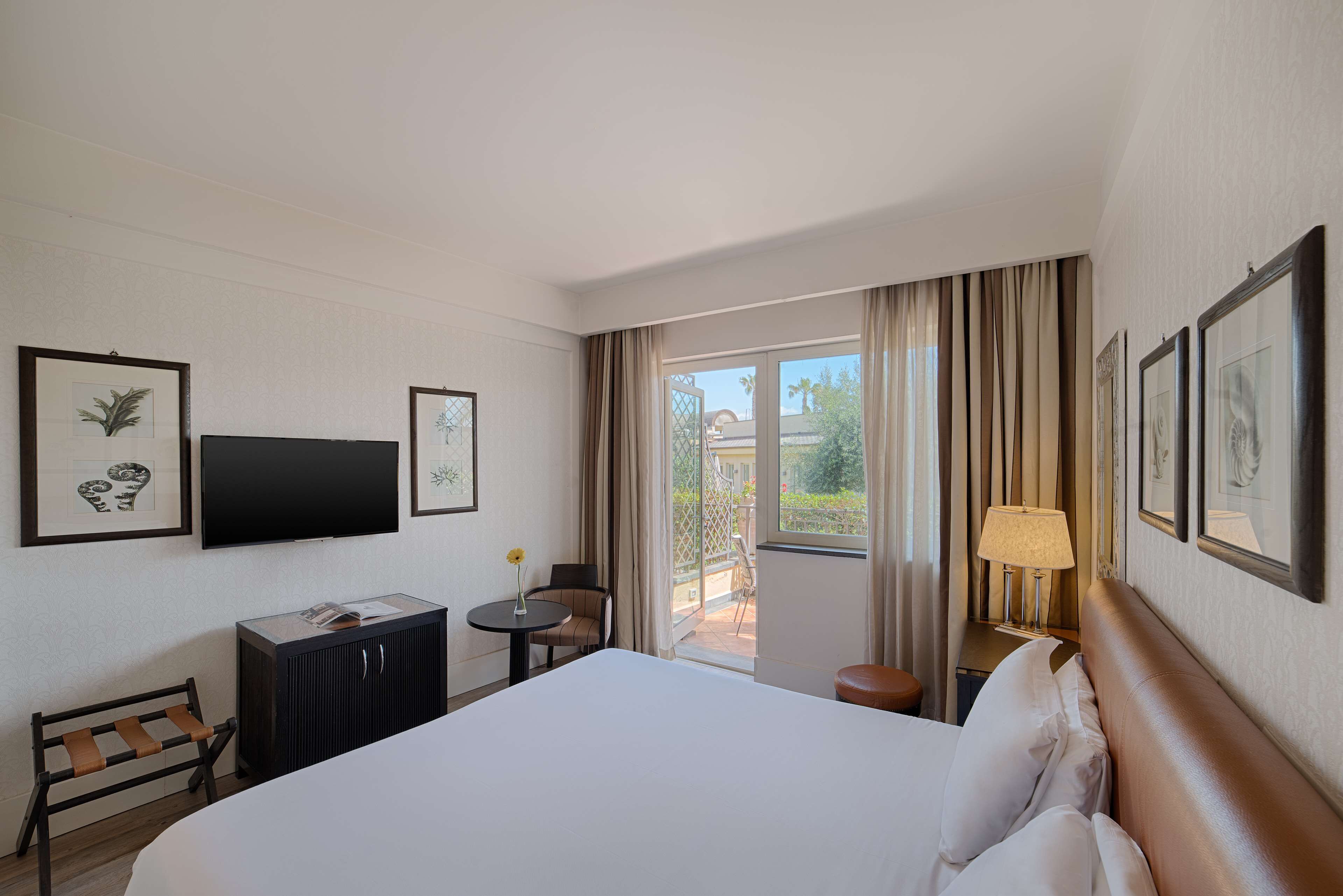 A hotel room with a large bed, a desk with a chair, a small table with a lamp, and a view of the outdoors.