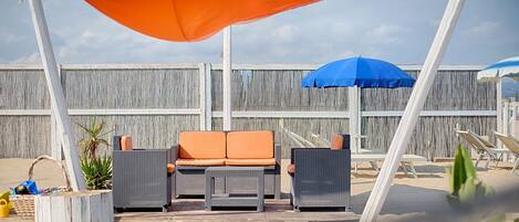 Une terrasse au bord de la plage, avec des meubles en osier, un parasol bleu et une clôture en bois en arrière-plan.