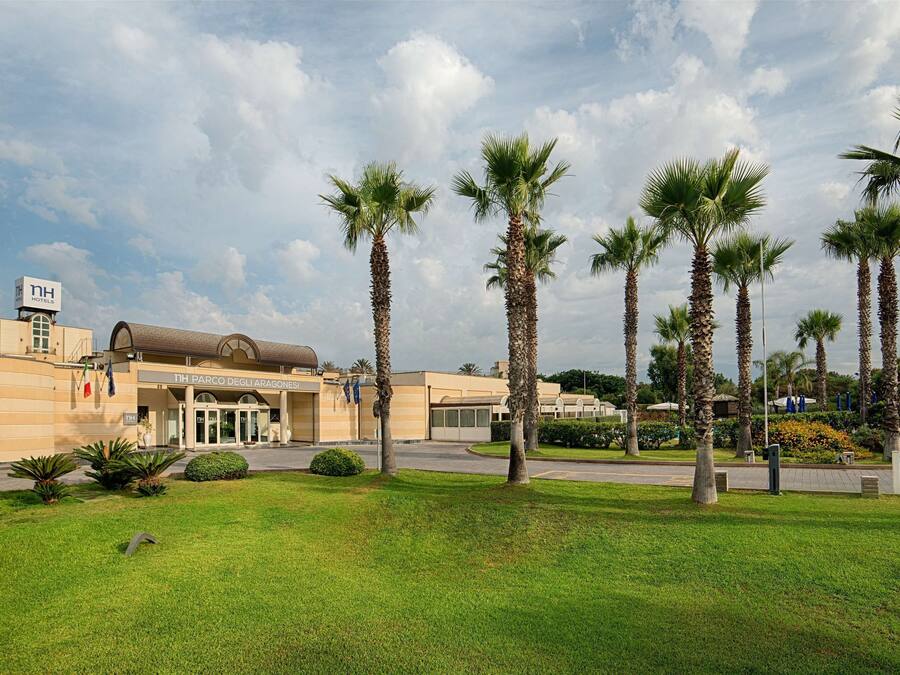 A hotel entrance with palm trees, a clear sky, and a sign for "1st Parco degli Aranci".