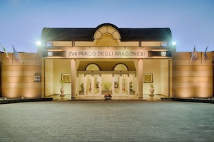 Exterior do hotel NH Parco degli Aragonesi, com entrada bem iluminada, plantas em vasos grandes e bandeiras de ambos os lados.