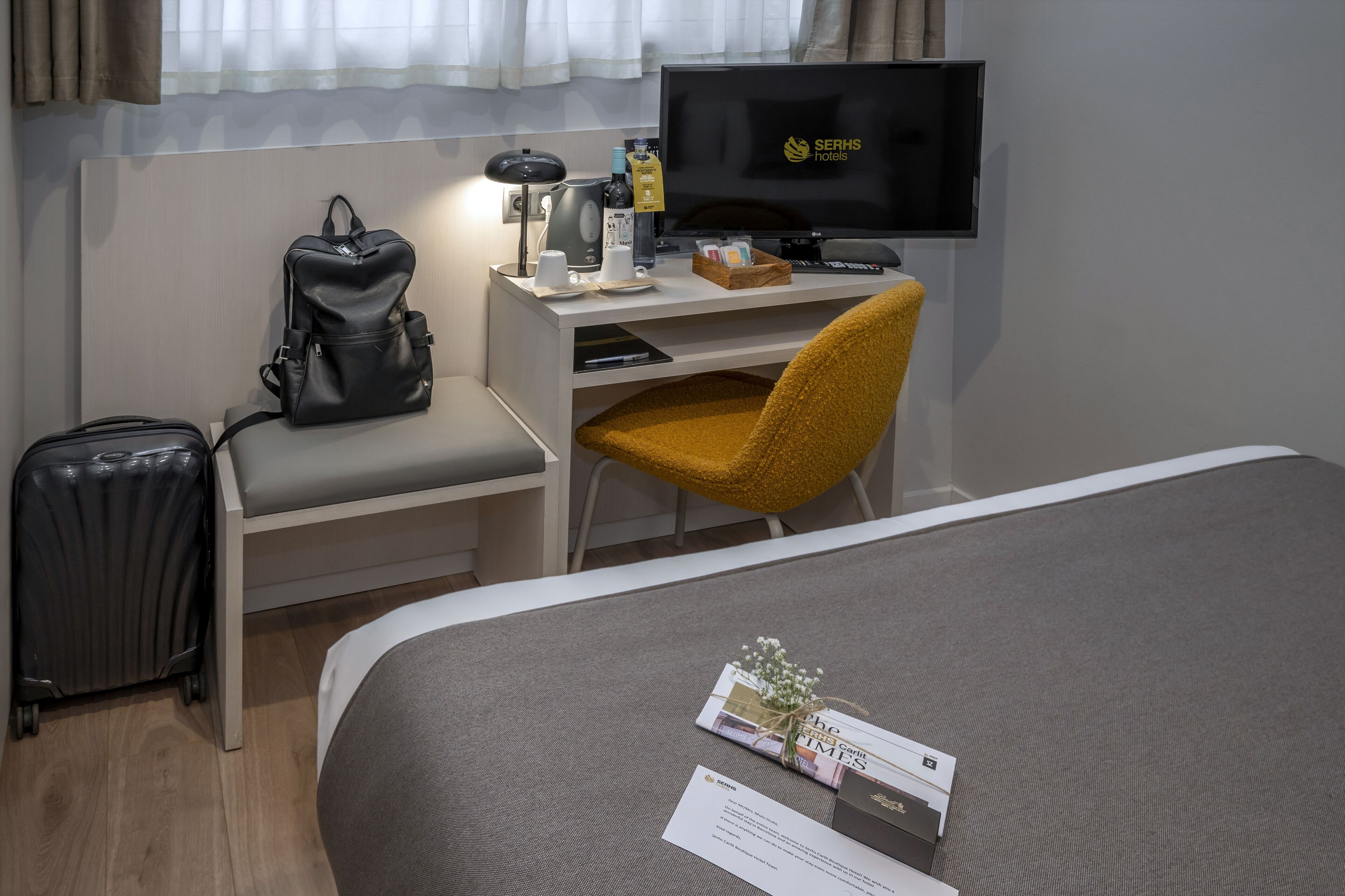 Una habitación de hotel con una cama, un escritorio con computadora, un televisor, una maleta y una silla.
