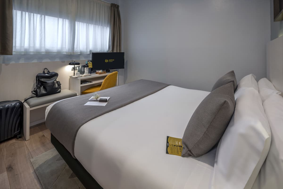A hotel room with a bed, a desk with a computer, a chair, a suitcase, and a window with curtains.