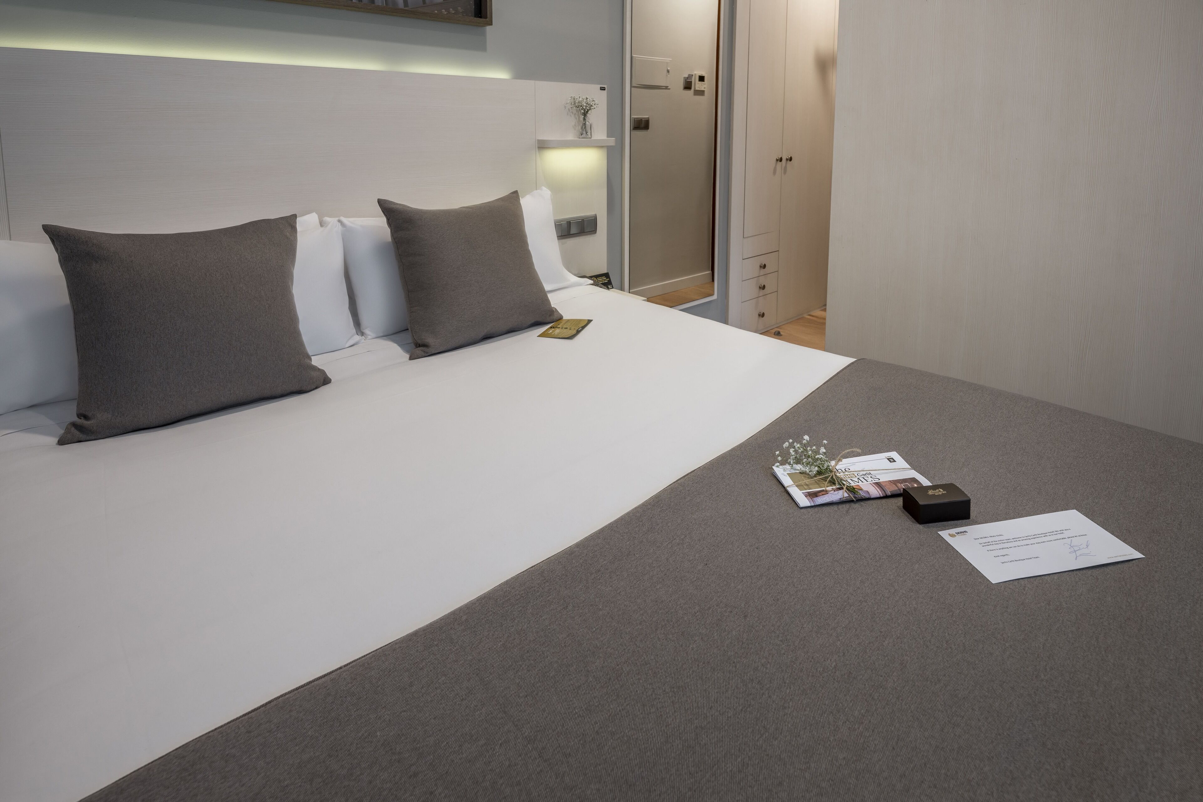 A neatly made bed with grey and white bedding, a bedside table with a book and a small box, and a door leading to a bathroom.