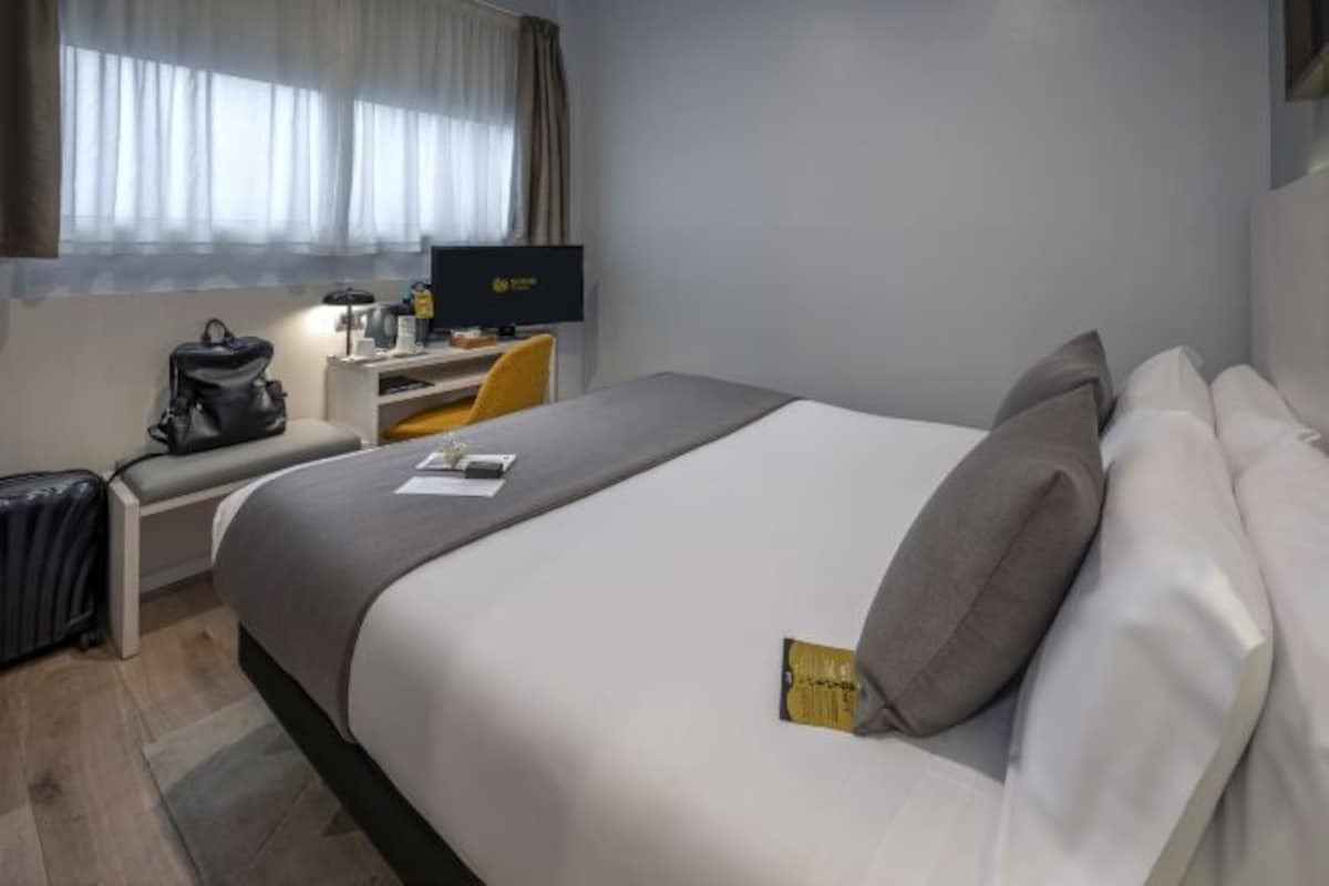 A hotel room with a bed, a desk with a computer, a chair, and a window with curtains.