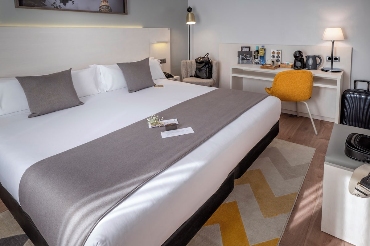 A hotel room with a large bed, a desk with a chair, a coffee maker, and a lamp.