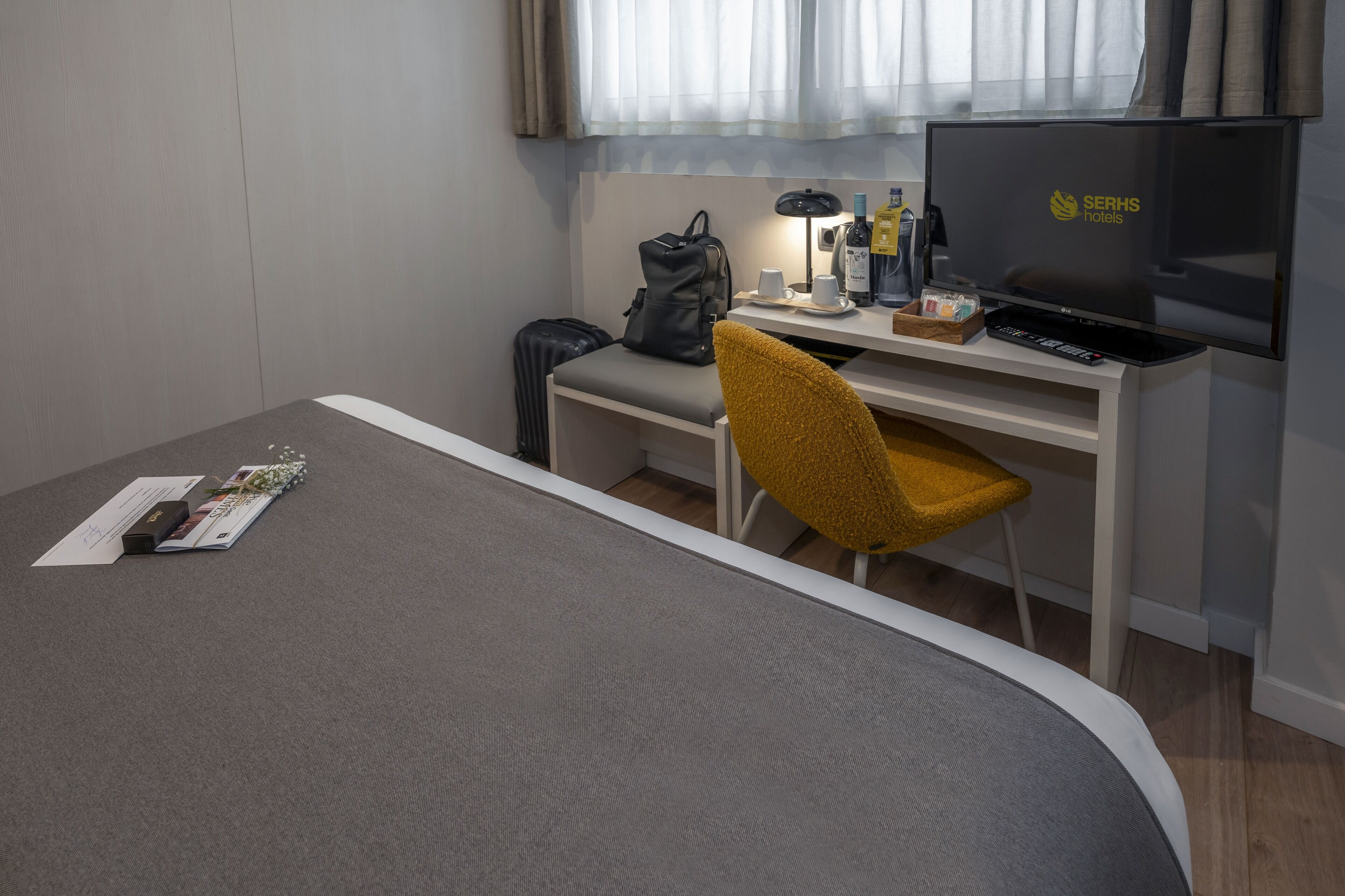 A hotel room with a bed, a desk with a TV, a chair, a lamp, and a window with curtains.