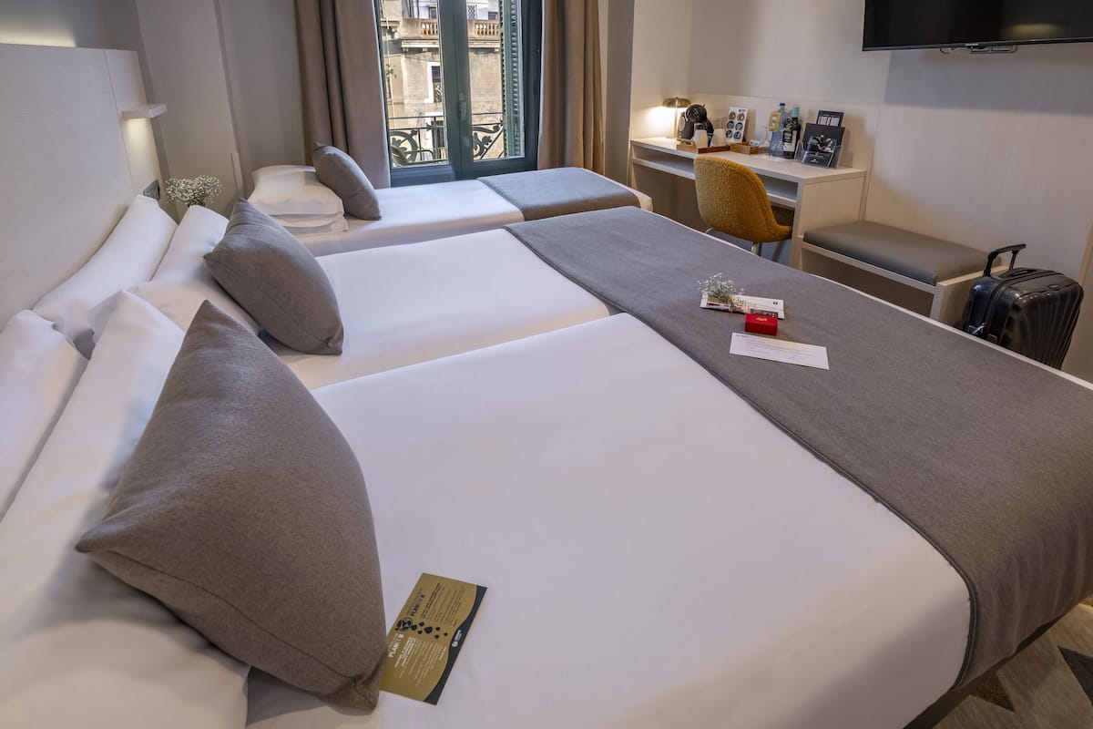 A hotel room with two beds, a desk, a chair, a TV, and a window with a view of buildings.