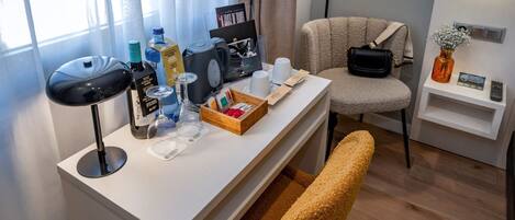 A well-lit hotel room with a white desk, a black desk lamp, a bottle of water, wine glasses, a coffee maker, and a chair.