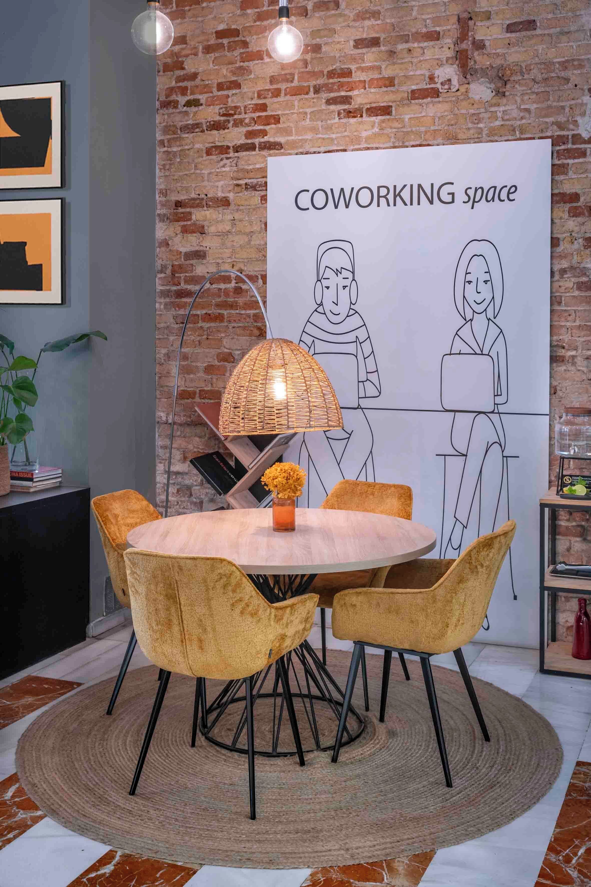 Um espaço de coworking com uma mesa redonda, quatro cadeiras, uma luminária e um cartaz na parede de tijolos.