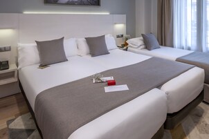 A hotel room with two beds, a bedside table with a phone, a small vase with flowers, and a window with curtains.