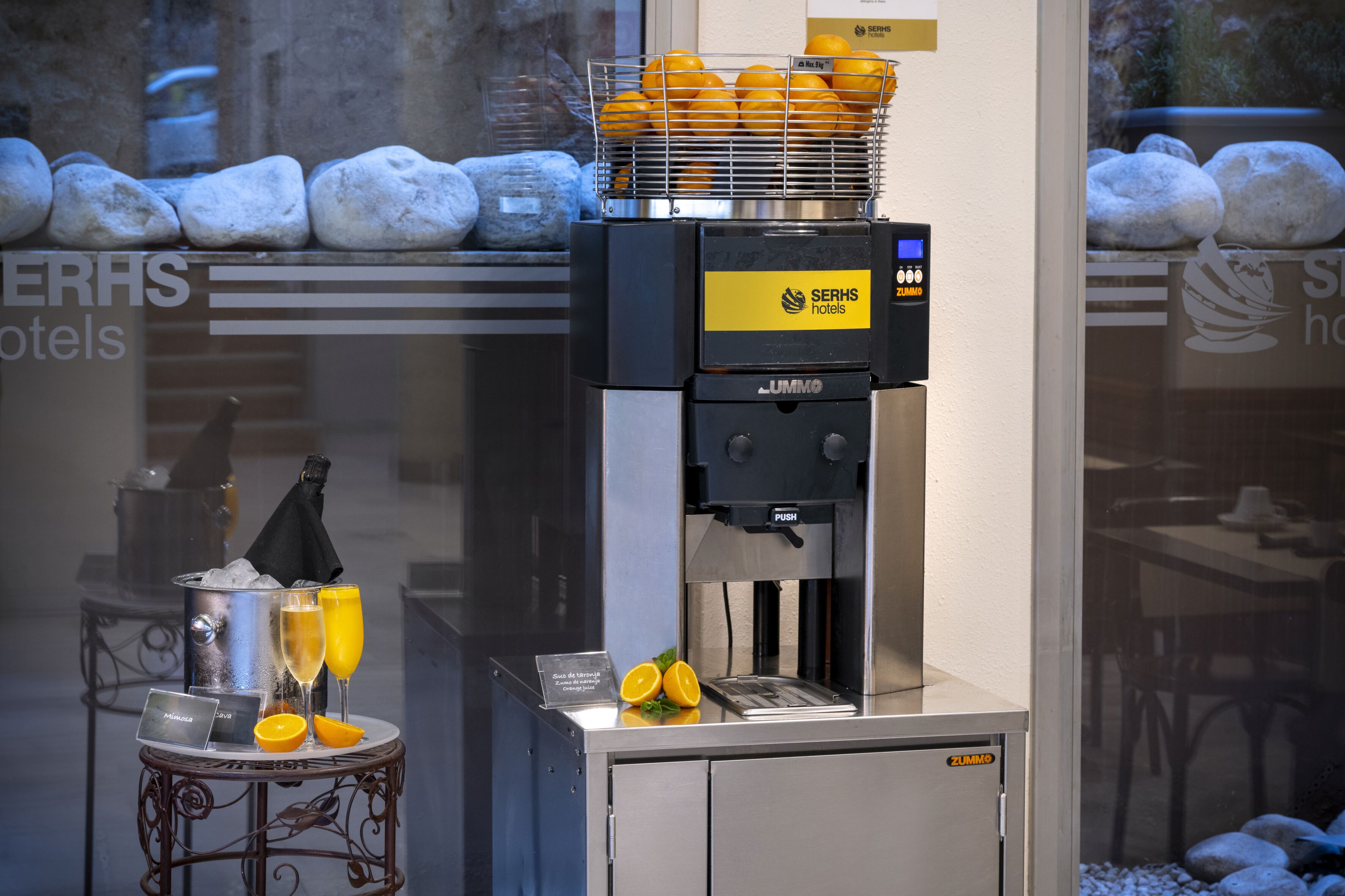 Eine Kaffeemaschine mit einer Orangenschale, einem Tablett mit zwei Gläsern Orangensaft und einem Schild für SerH's Hotels.