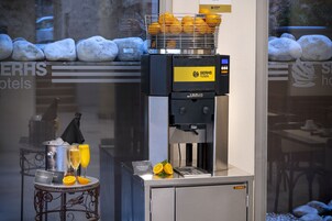 A coffee machine with a basket of oranges, a tray with two glasses of orange juice, and a sign for SerH's hotels.