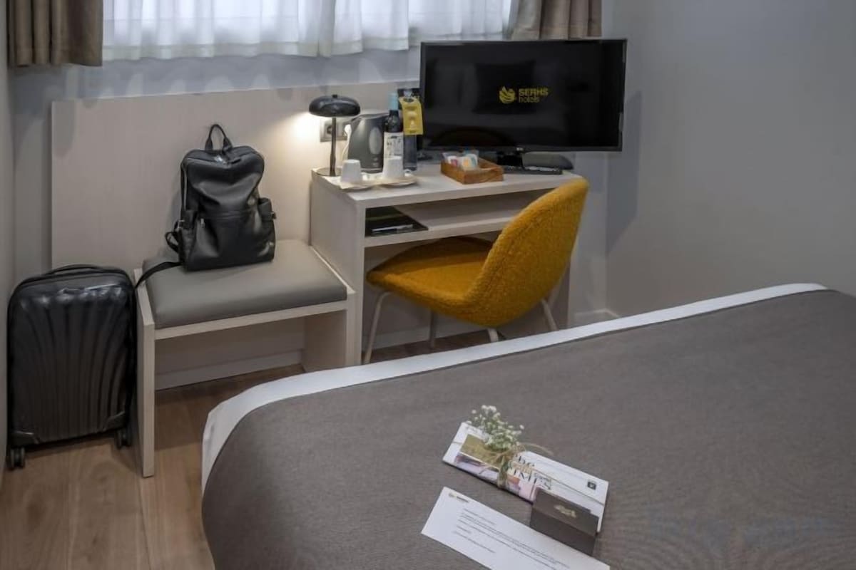 Ein Hotelzimmer mit einem Bett, einem Schreibtisch mit Computer, einem Stuhl, einem Koffer und einem Fernseher.