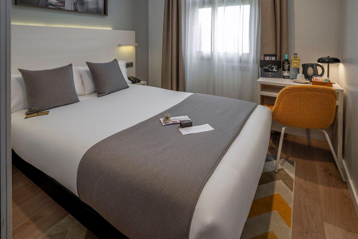 A neatly made bed with a grey blanket, a small bedside table with a lamp, and a chair with a yellow cushion.