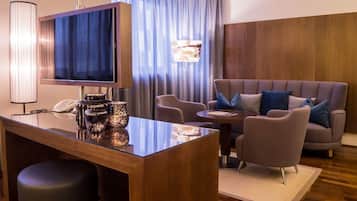 A hotel room with a wooden desk, a flat-screen TV, a sofa with cushions, and a large window with curtains.