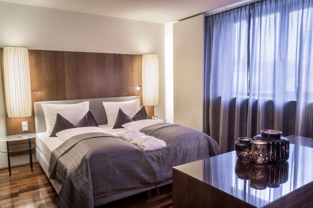 A modern hotel room with a large bed, two bedside lamps, a glass table with decorative items, and a view of the outside through sheer curtains.