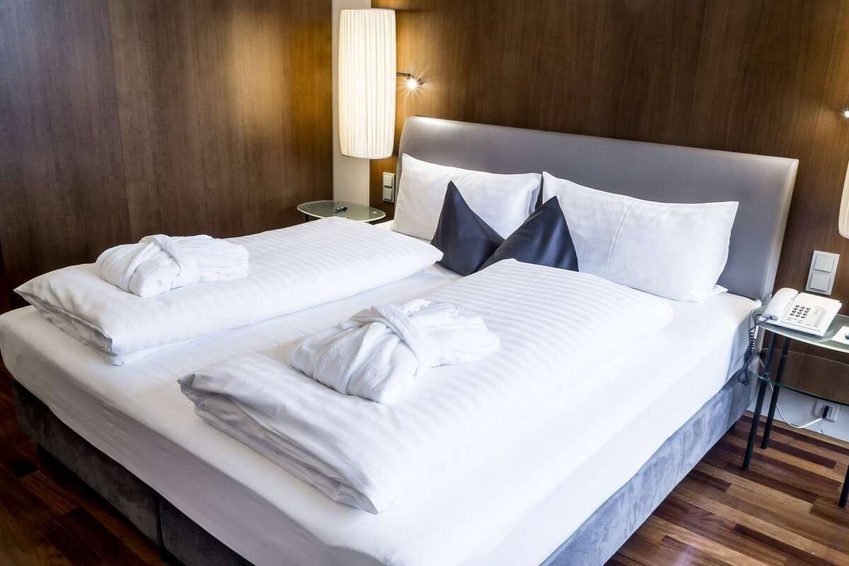 A double bed with white linens and two towels, a bedside lamp, a phone, and a wooden wall.