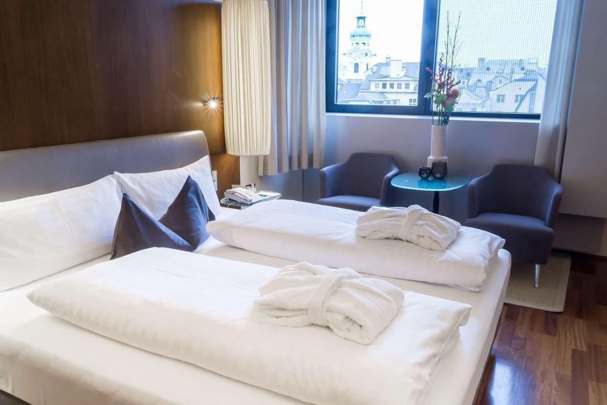 A hotel room with two beds, a small table with a vase, and a view of the city through a large window.