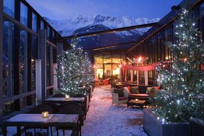 A snow-covered outdoor seating area with two Christmas trees, tables, and chairs, overlooking a mountain range. - The Penz Hotel (Innsbruck)
